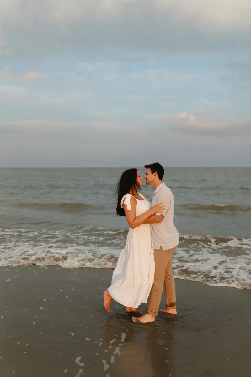 A couple embraces and shares a kiss while standing in the shallow waves of a beach at sunset, with calm waters and soft clouds in the background.