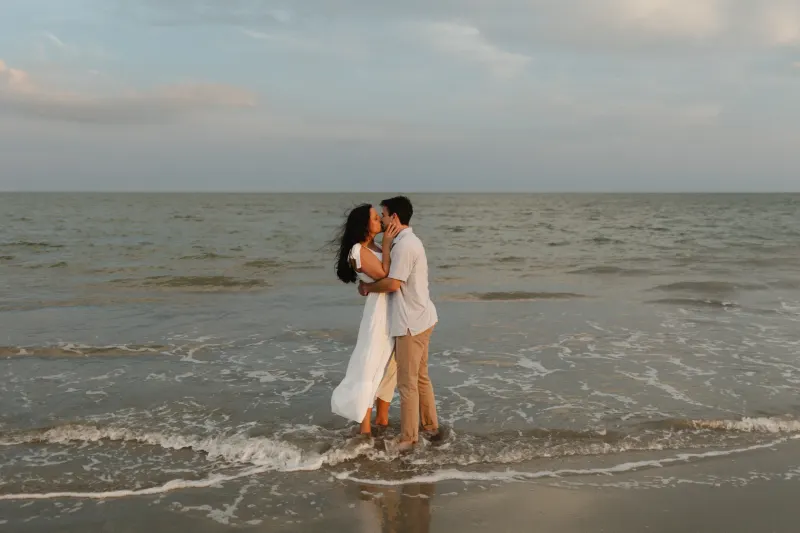 A couple in white clothing embraces and kisses in the shallow ocean waves during what appears to be a wedding or romantic photoshoot at sunset.