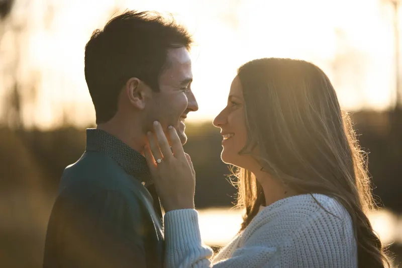 A man and woman face each other intimately outdoors during golden hour, with the woman's hand gently touching the man's face as they smile at each other near a body of water.