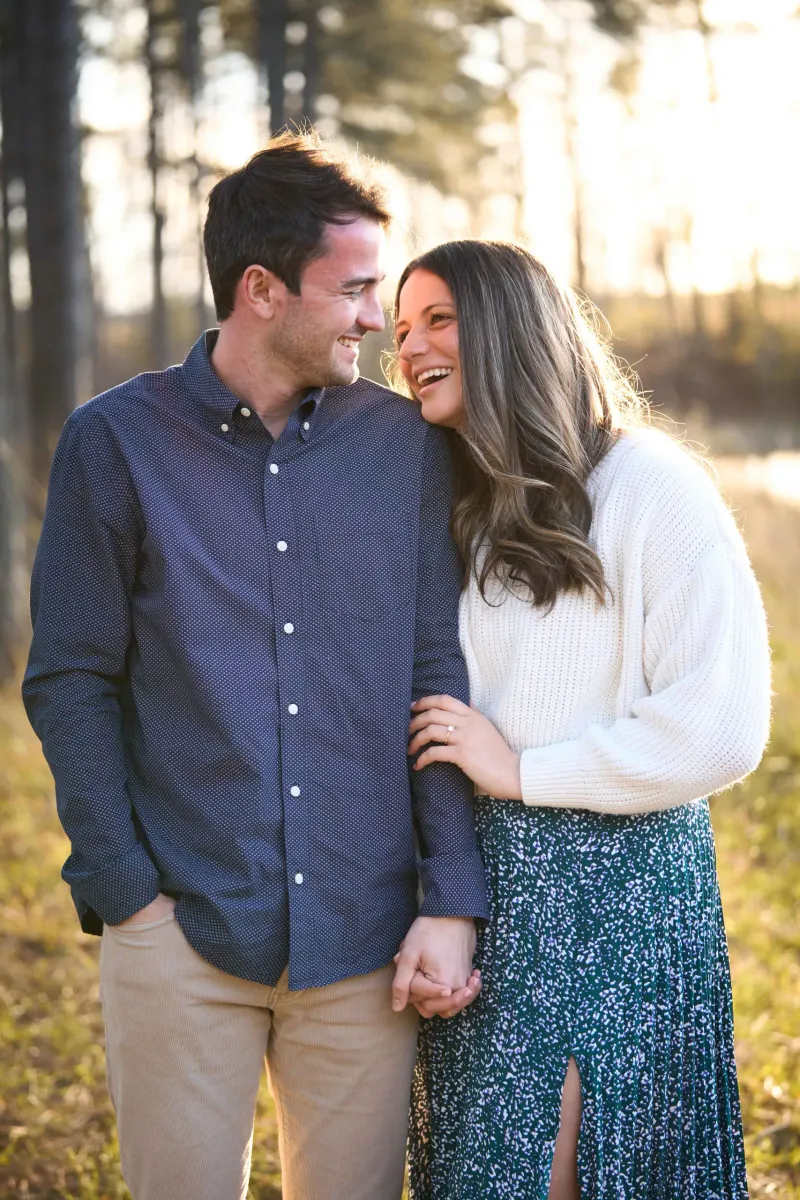 A smiling couple holds hands while standing together in a sunlit forest clearing, with tall pine trees and golden hour lighting in the background.